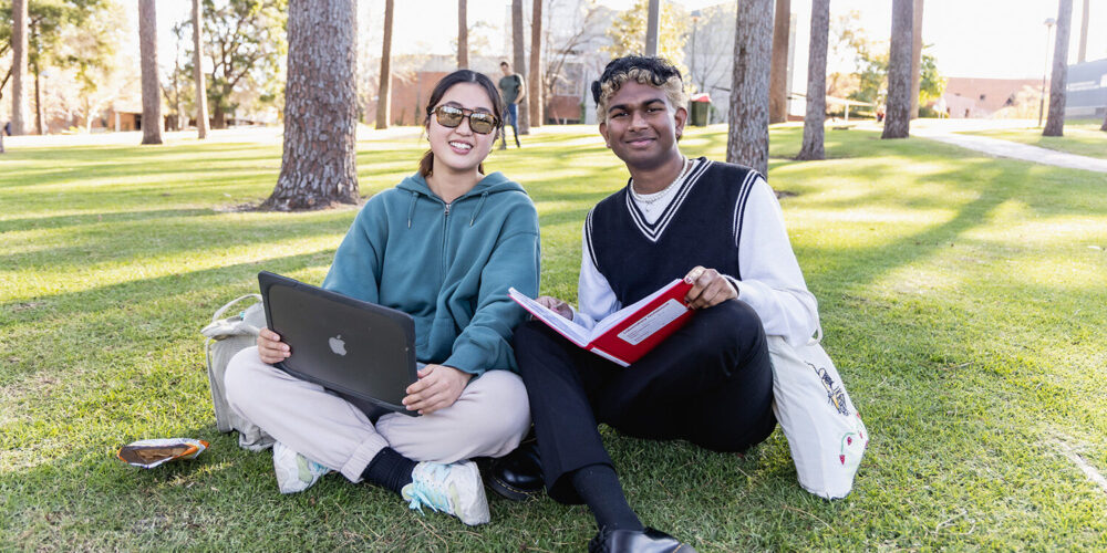 2 students sitting on the grass, smiling holding laptops and textbooks.