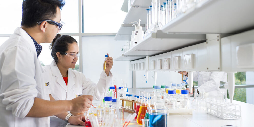 Two students in lab coats looking at colourful chemical glassware