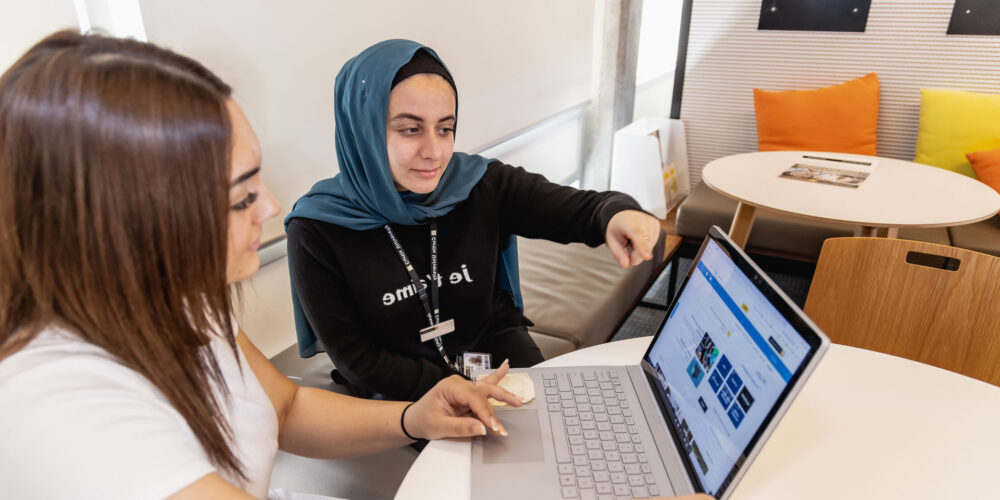 A student points to a laptop screen while another student watches.